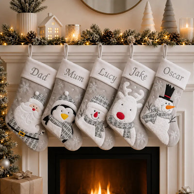 Five personalized Christmas stockings hang from a fireplace mantel decorated with pine branches, fairy lights, candles, and a small tree. The stockings are light gray with white cuffs and embroidered holiday characters: Santa, penguin, snowman, reindeer, and snowman.