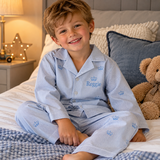 A smiling young boy sits on a bed wearing light blue and white striped pajamas with a small crown pattern and the name 'Reggie' embroidered on the chest. The bedroom setting includes a bedside table, lamp, star-shaped night light, pillows, and a teddy bear.