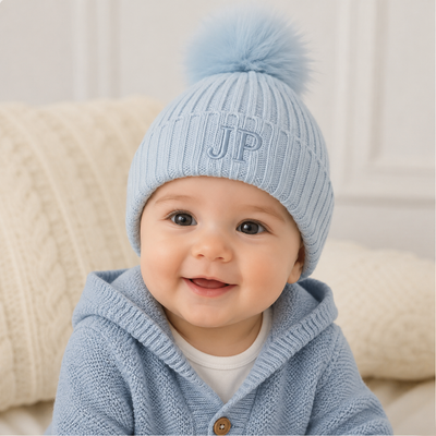 Baby boy wearing a baby blue knitted pom hat personalised with the initials JP in an embroidered font on the front of the hat.