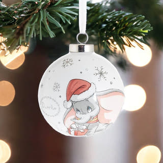 A round white Christmas ornament featuring an illustration of Dumbo wearing a Santa hat, hanging from a green pine branch with warm bokeh lights in the background.