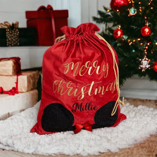 A red Christmas gift bag with gold 'Merry Christmas Millie' lettering and black Mickey-shaped feet, placed on a white fluffy rug in front of a decorated Christmas tree and wrapped gifts.