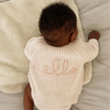 Baby wearing a beige knitted outfit with 'hello' on it, lying on a soft surface.
