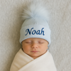 Newborn baby wearing a light blue knit hat with a pom-pom and 'Noah' embroidered on it, wrapped in a white blanket.