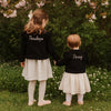 Two young girls holding hands with 'Penelope' and 'Posey' on their black shirts, standing in a garden.