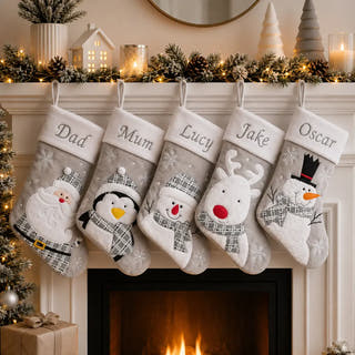 Five personalized Christmas stockings hang from a fireplace mantel decorated with pine branches, fairy lights, candles, and a small tree. The stockings are light gray with white cuffs and embroidered holiday characters: Santa, penguin, snowman, reindeer, and snowman.