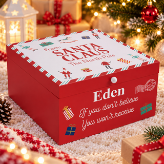 A red personalized Santa gift box with a white top lid, decorated with holiday illustrations and the text 'From: Santa Claus The North Pole' and 'Eden If you don't believe You won't receive'. The box is styled on a white textured rug with Christmas lights, pinecones, and wrapped presents in the background.