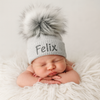 Newborn baby wearing a gray knit hat with a fluffy pom-pom, lying on a white fluffy surface.