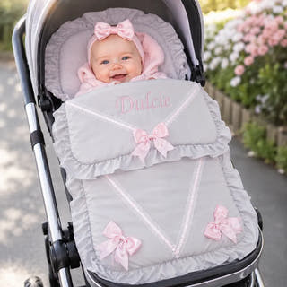 A baby wearing a pink bow headband is seated in a stroller, wrapped in a light gray footmuff with pink bows and the name 'Dulcie' embroidered on the front. The stroller is shown outdoors with blurred flowers in the background.