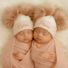 Two sleeping babies wrapped in pink blankets, wearing white hats with fluffy pom-poms and embroidered names.