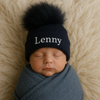 Newborn baby wrapped in blue fabric and wearing a black knit hat with a pom-pom on a beige background