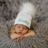 Newborn baby wearing a white knit hat with a large pom-pom and embroidered name 'Marley' on a soft, textured surface.