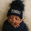 Newborn baby wearing a navy blue knit hat with a pom-pom and 'THEO' embroidered on it, wrapped in dark blue fabric against a beige textured background.