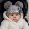 Baby wearing a gray knit hat with two pom-poms and the name 'CODY' in white text, sitting in a stroller.