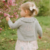 Child wearing a gray cardigan with 'Abby' embroidered on it, standing in a garden with pink flowers.