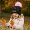 Child wearing a black beanie with pink pom-pom and 'Hallie' embroidered, holding autumn leaves outdoors.