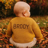Baby wearing a yellow knitted sweater with 'BRODY' on the back, sitting in an autumn setting.