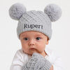 Baby wearing a gray knit hat with pom-poms and the name 'Rupert' on a white background