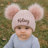 Baby wearing a pink knit hat with fluffy pom-poms and 'Kelsey' embroidered on it, standing outdoors.