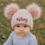 Baby wearing a pink knit hat with fluffy pom-poms and 'Kelsey' embroidered on it, standing outdoors.