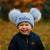 Child wearing a blue knit hat with pom-poms and 'Kallum' embroidered, standing outdoors.