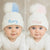 Two babies wearing white beanies with colorful pom-poms, one named 'Rory' and the other 'Thea'.