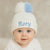 Baby wearing a white knit hat with a blue pom-pom and embroidered name 'Rory' on a beige background