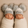 Two newborn babies wearing personalized hats with fluffy pom-poms on a beige background.