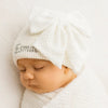 Newborn baby wearing a white knit hat with a bow, on a light background