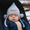 Baby wearing a knitted hat with 'Ryan' embroidered on it, sitting in a stroller outdoors.