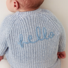 Baby wearing a blue knitted cardigan with 'hello' embroidery on a plain background