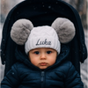 Baby wearing a white knit hat with gray pom-poms and 'Luka' embroidered, sitting in a stroller.