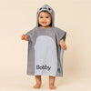 Child wearing a gray shark-themed poncho with 'Bobby' printed on it against a beige background
