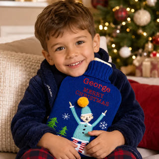 A young boy smiling while hugging a blue knitted Christmas sweater with 'George Merry Christmas' embroidered in red and a snowman design. He is wearing a navy fleece jacket and plaid pajamas, with a decorated Christmas tree in the background.