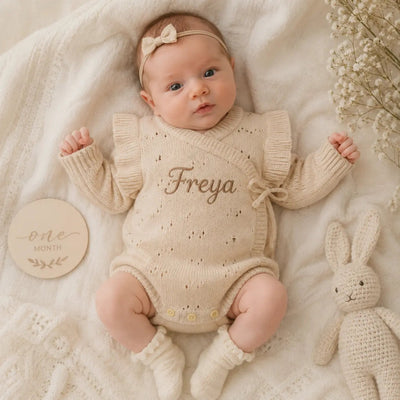 Baby in a beige knitted outfit with 'Freya' embroidered, lying on a soft surface with a wooden plaque and toy.